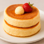 Dos pancakes japoneses esponjosos apilados en un plato blanco, coronados con una porción de mantequilla y una fresa, con sirope brillante deslizándose por la superficie.
