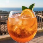 Copa de Aperol Spritz naranja brillante, lleno de hielo y adornado con una rodaja de naranja, en un ambiente soleado de terraza.
